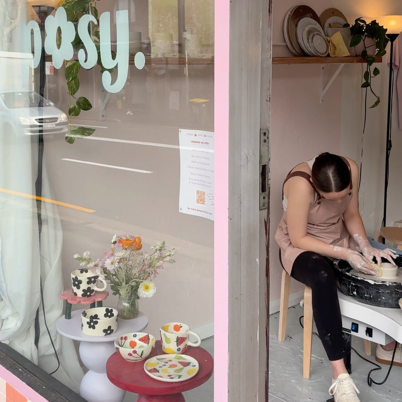 Person sitting on a pottery wheel stool inside a store with a pink and white interior, looking out the window at a street view.